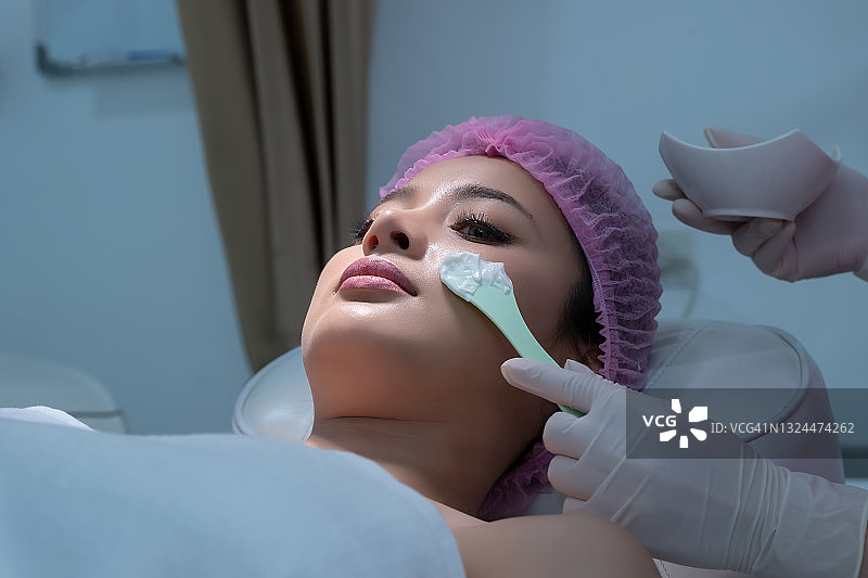 临床保健，美容师或皮肤科医生把白色霜刷在美丽的女人的脸上在美容诊所。图片素材