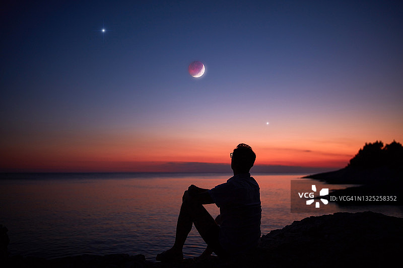 一个人的剪影看着月亮和星星越过海洋的地平线。图片素材
