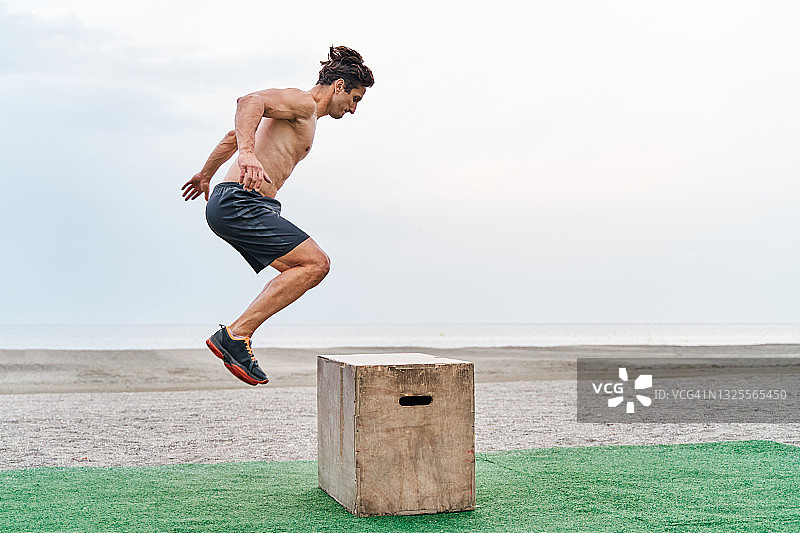 一个年轻的肌肉发达的年轻男子在沙滩上训练赤膊跳上箱子。图片素材