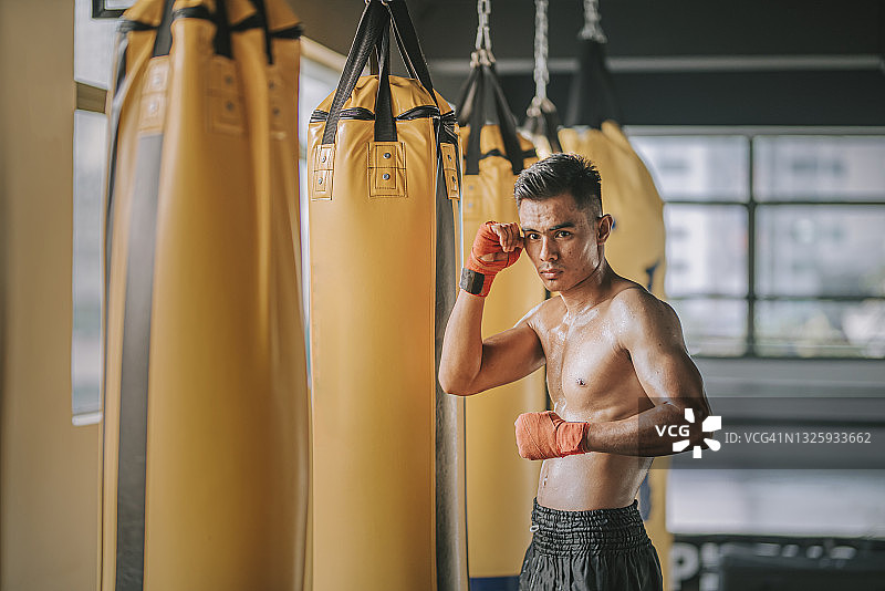 亚洲马来人泰拳男运动员肖像看着相机旁边一排拳击袋在健身房图片素材