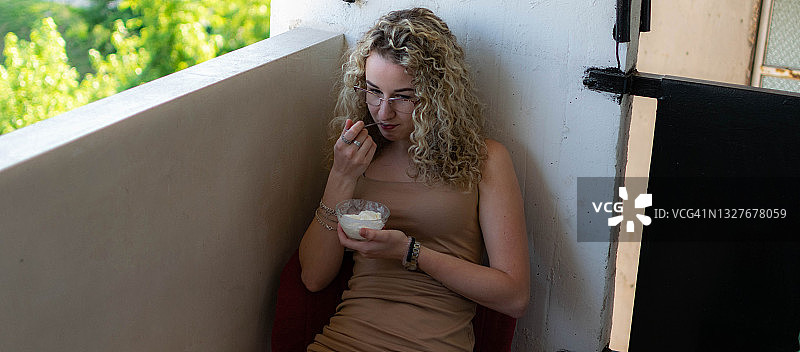 年轻的卷发女子坐在家里，享受着吃冰淇淋，享受着新鲜空气，吃着冰激凌在她的阳台上，同时使用着笔记本电脑。图片素材