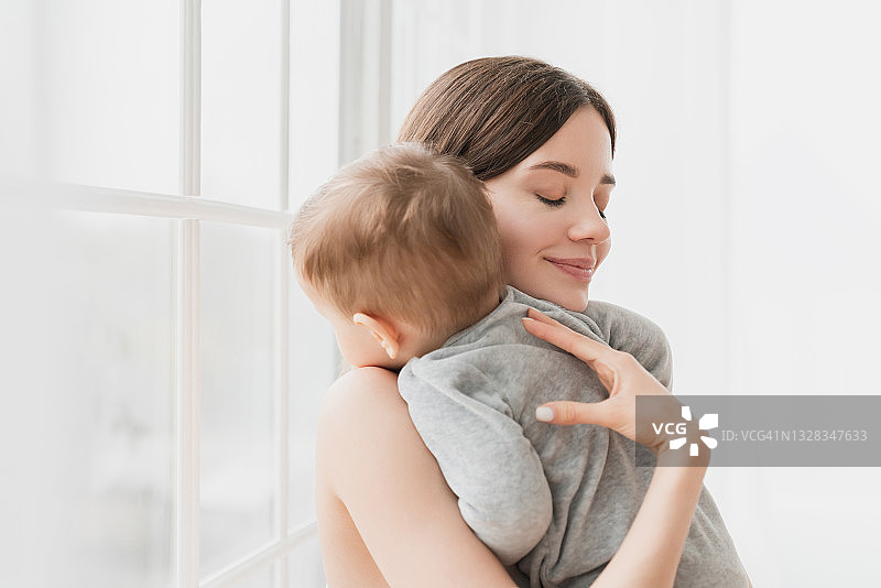 年轻的单身母亲抱着她年幼的儿子女儿蹒跚学步的婴儿刚出生的婴儿哄着她的孩子睡觉，午睡和睡觉时间。母亲和儿童保育概念图片素材
