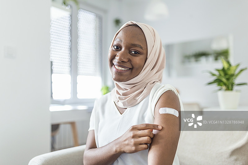 一名非洲穆斯林妇女在接种疫苗后微笑的肖像。妇女按住她的衬衫袖子，并显示她的手臂与绷带接受疫苗接种后。图片素材