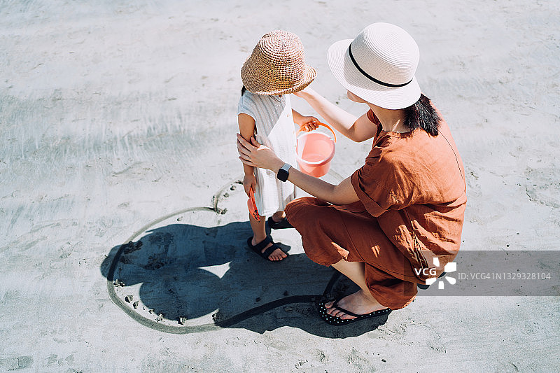 在一个阳光明媚的夏日，年轻的亚洲母亲和小女儿在沙滩上画着一颗心，一起玩耍，可爱的女儿站在心里。家庭的爱与关怀图片素材