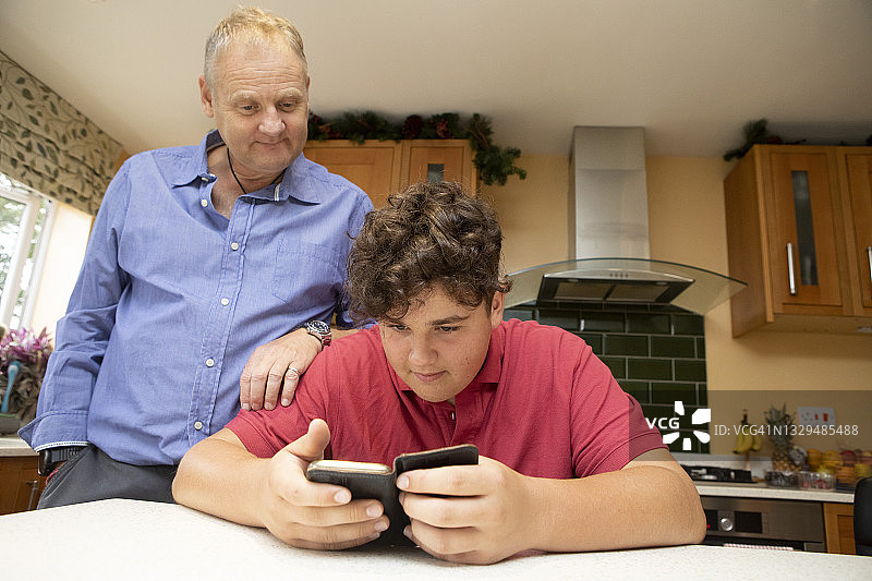 成熟的男人和十几岁的男孩一起看手机在一个家庭厨房图片素材