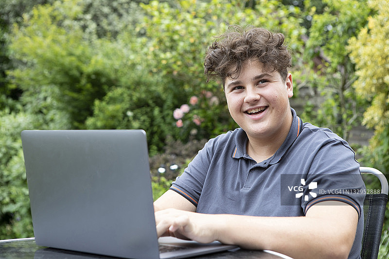 一个十几岁的男孩坐在花园的桌子旁，用着笔记本电脑，看着旁边，微笑着图片素材