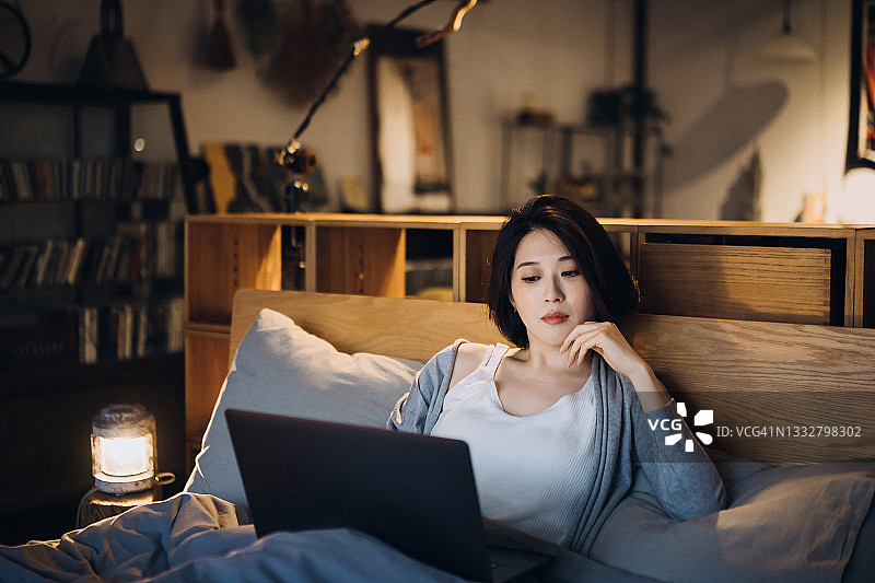 美丽的年轻亚洲女人躺在床上，在睡觉前上网和在笔记本电脑上购物，在舒适的家里度过一个寒冷的夜晚图片素材