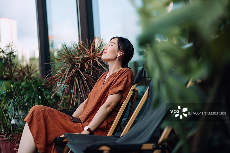 年轻的亚洲妇女与眼睛闭上享受新鲜空气，同时放松在阳台躺椅在早上，被美丽的室内植物包围。生活方式和健康概念图片素材