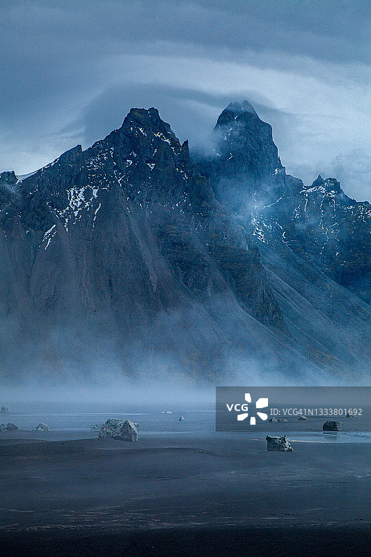 宽镜头的黑色沙滩与雾升起的海洋和戏剧性的山脉和云彩填充天空的背景。图片素材