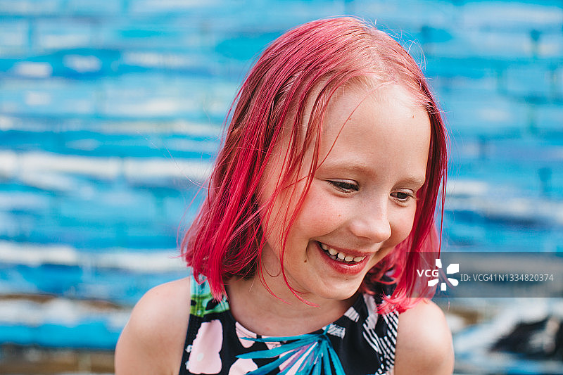 一个穿着游泳衣、一头粉红色头发的女孩，在一堵漆成海蓝色波浪的砖墙的背景下微笑、做鬼脸、翻白眼，玩得很开心。图片素材