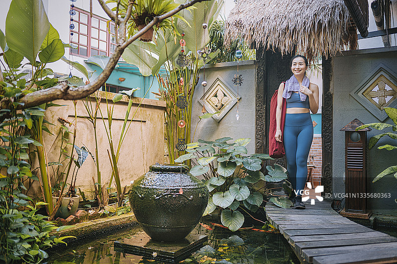 亚洲的中国美女穿着运动服和瑜伽垫穿过木制人行桥上锦鲤池塘花园巴厘岛风格图片素材