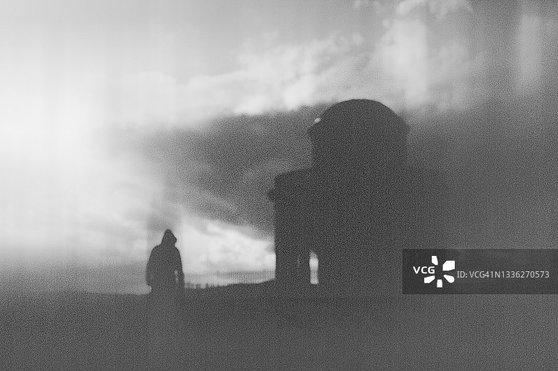 一个黑暗的幻想概念。一个孤独的戴着兜帽的人，站在一座古老的历史建筑旁边。模糊抽象的编辑。图片素材