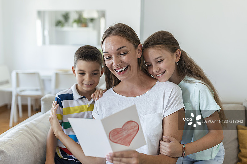 快乐的年轻母亲和孩子们坐在沙发上，拥抱着，读着家里寄来的明信片上的节日祝福和祝贺图片素材