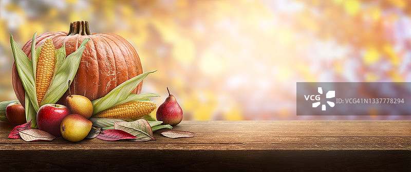 一张由南瓜、苹果、梨子和玉米芯组成的感恩节庆祝桌，背景是秋天日落的树叶。图片素材