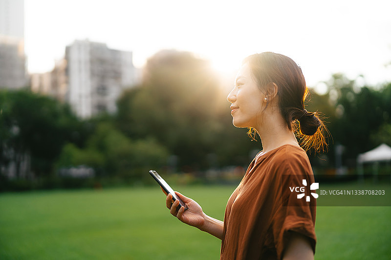 美丽微笑的年轻亚洲女人的侧面正在看风景，在公园的阳光下使用智能手机放松。享受一个无忧无虑、轻松的下午。生活方式和技术。与自然的联系图片素材