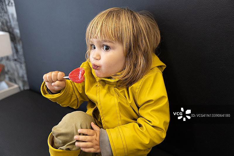 穿着黄色雨衣的小孩坐在沙发上吃着甜棒棒糖。图片素材
