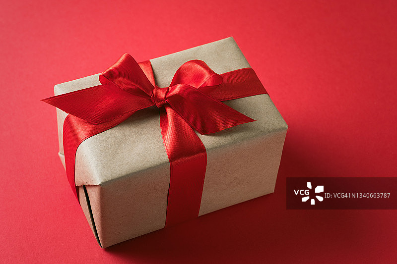 一个惊喜的包裹，用牛皮纸包着，用鲜亮的丝带系在红色的背景上。在节日前夜送礼物。圣诞快乐，情人节概念。图片素材