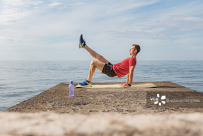 一个全神贯注的年轻人站在海边的运动垫上做着腹肌锻炼图片素材