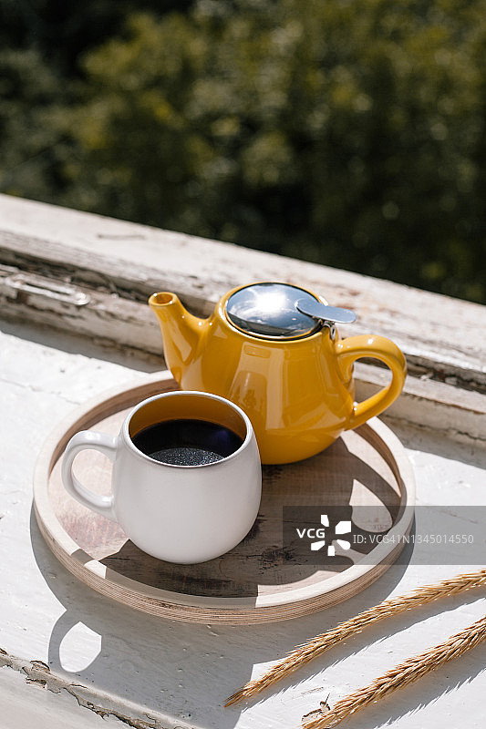 在明亮的阳光下，一杯茶和木制托盘上的黄色水壶。白色面料背景。早上的概念。图片素材