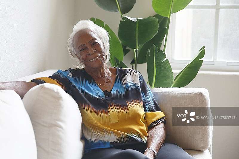 在明亮的客厅里，一位白发苍苍的非裔美国老年妇女穿着色彩鲜艳的上衣，愉快地坐在那里。图片素材