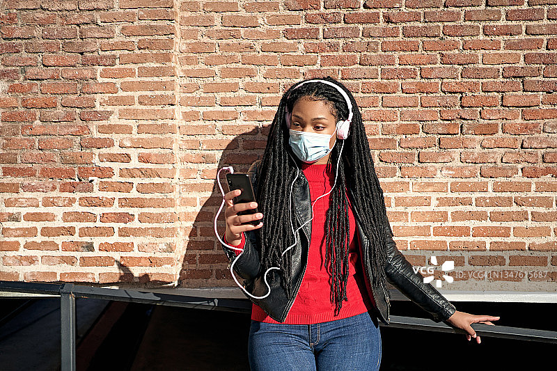 一名非裔美国妇女的肖像戴着口罩，拿着手机在城市的街道上听音乐。头发被编成了脏辫。新常态的概念图片素材