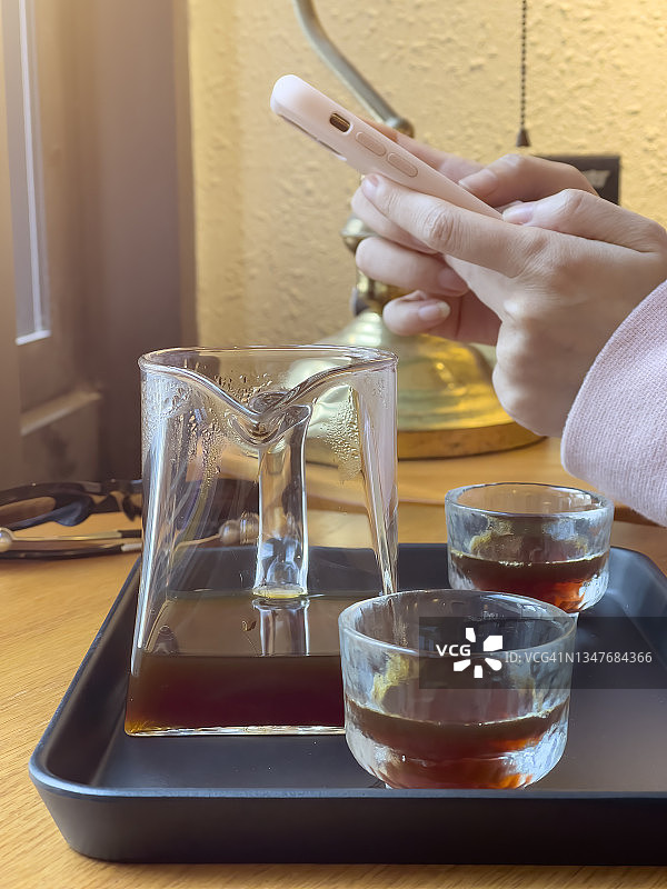 女人用手机，玻璃咖啡杯和咖啡壶都放在桌子前面图片素材