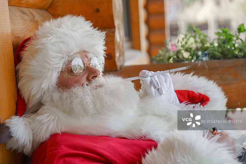 这是一个阳光明媚的夏日，圣诞老人悠闲地坐在他木屋的阳台上的椅子上，享受着白色烟嘴里的香烟图片素材