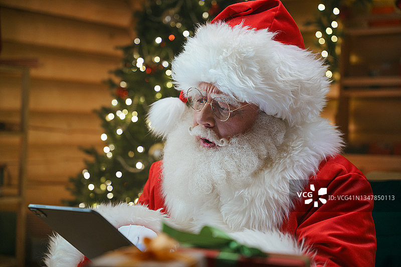 圣诞老人正在数码平板电脑上读着什么，他坐在一间小屋里，装饰着圣诞节的装饰图片素材