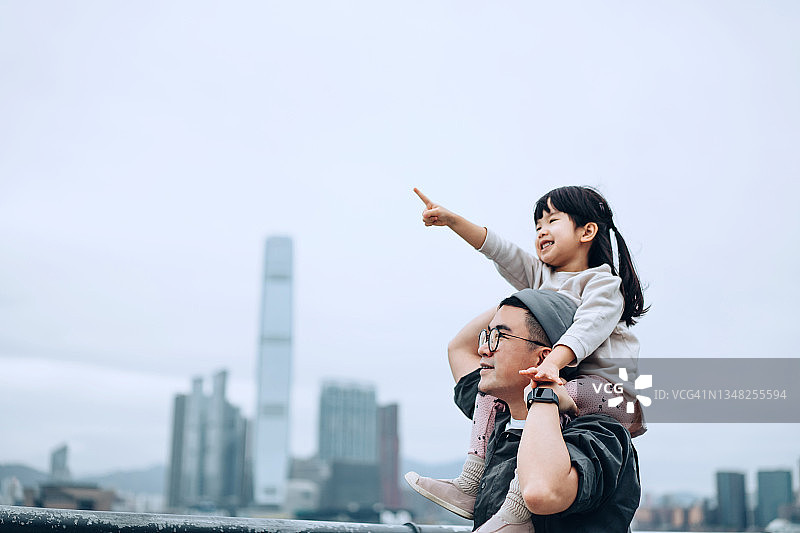 年轻的亚洲父亲带着小女儿骑在维多利亚港的海滨长廊上，小女孩指着远方，眺望着美丽的城市天际线图片素材