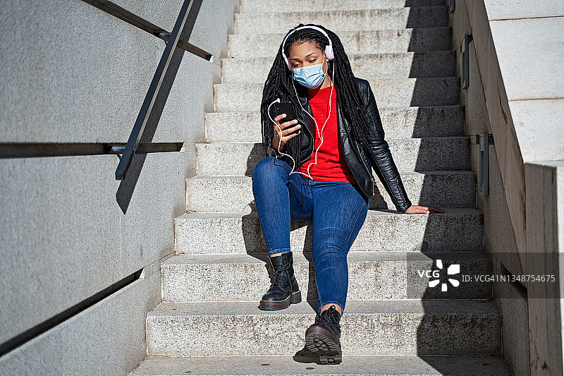 一名非裔美国妇女的肖像戴着口罩，拿着手机在城市的楼梯上听音乐。头发被编成了脏辫。新常态的概念图片素材