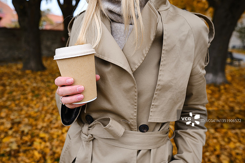 女人拿着一个工艺纸杯，里面放着咖啡或茶，在秋天的公园里散步。喝咖啡的时间。一次性纸杯。带走。热饮料。空白的空间图片素材