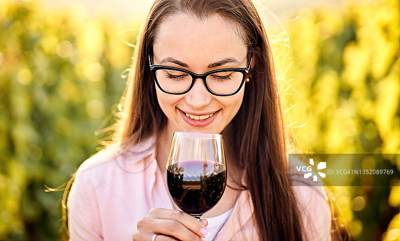 一个年轻的，千禧一代的女人的肖像，在葡萄园的户外拿着和闻着一杯有机生物红葡萄酒-葡萄种植和品酒的概念图片素材