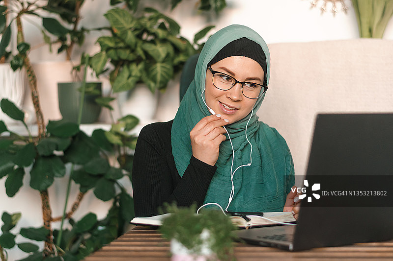 积极的年轻穆斯林妇女戴着头巾，戴着耳机，坐在一个有大量室内植物的舒适房间里使用笔记本电脑。业务、自由电子学习的概念,不同的人。图片素材