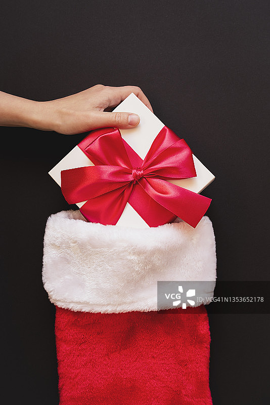 女人的手把一个带着红色蝴蝶结的白色礼物放在红色圣诞老人的靴子里。图片素材