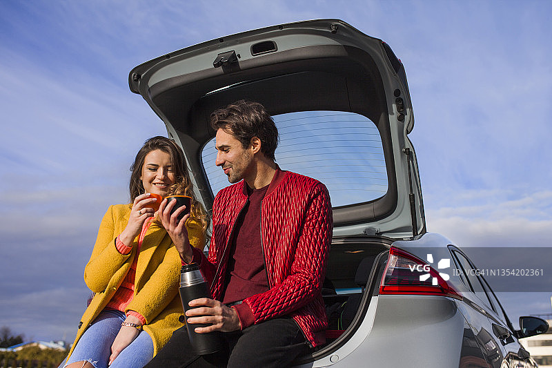 一对年轻快乐的夫妇坐在汽车后备箱里喝茶图片素材