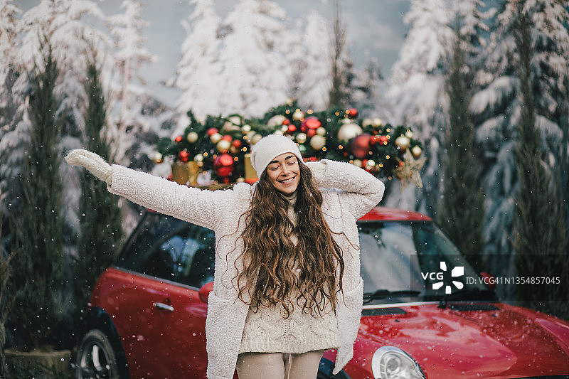 快乐的美女穿着针织毛衣，戴着羊毛帽，站在红色的汽车旁边，圣诞树在头顶，下着雪花。图片素材