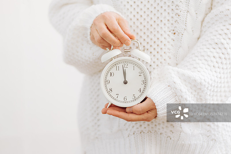 一个穿白色毛衣的女孩手里拿着一个象征新年的闹钟，箭头指向午夜12点。假期概念及新年图片素材