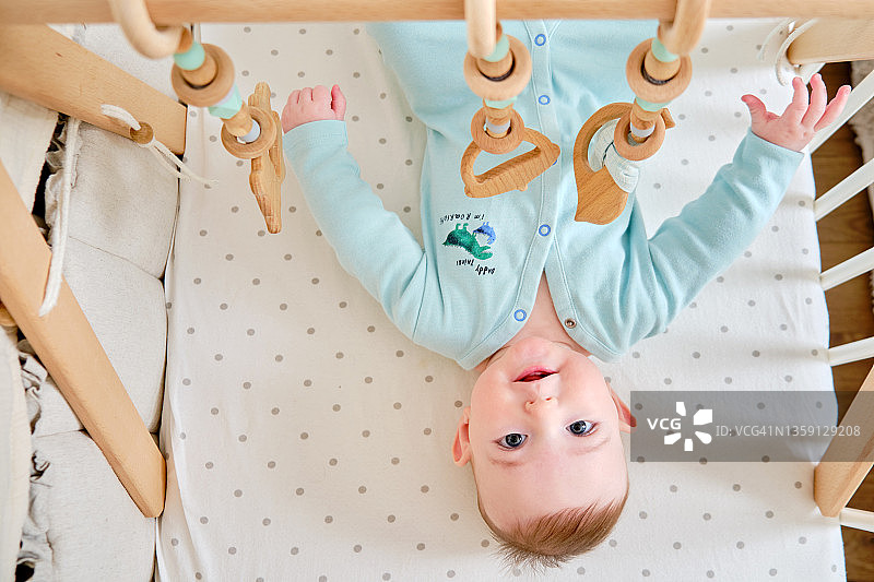 快乐的婴儿躺在床上玩着木制的吊玩具。6个月大的婴儿床里一个穿着绿松石衣服笑得滑稽的孩子图片素材