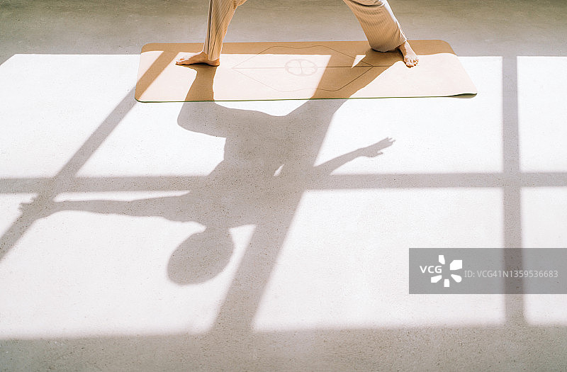 一个短发女人的影子在窗前的瑜伽垫上做着战士式的瑜伽。放松练习的概念图片素材