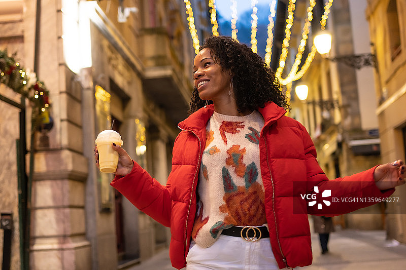 刻画黑人少数民族妇女冬天在城市的夜晚。喝着外卖橙汁在灯火通明的街道上漫步图片素材