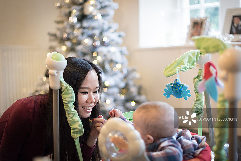 在英国苏格兰边境梅尔罗斯的一所房子里，一名亚洲妇女微笑着和一个装在步行玩具里的婴儿玩耍，窗户旁有一棵白色的圣诞树图片素材
