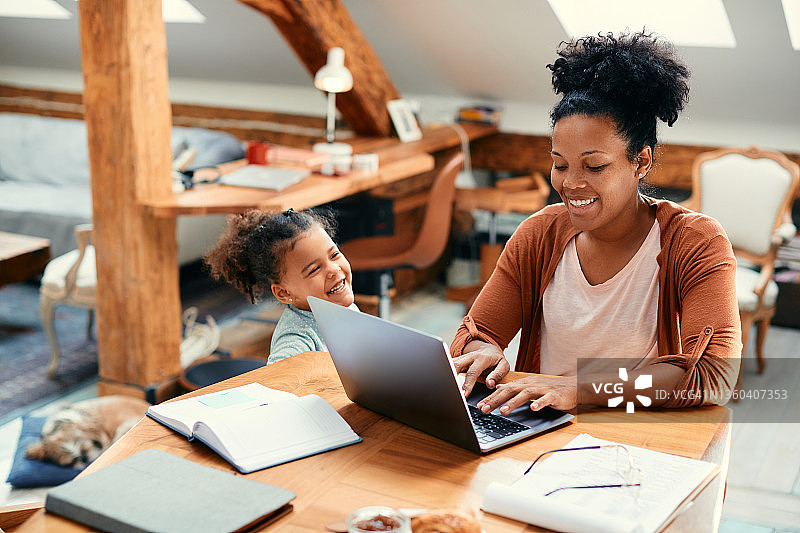 快乐的黑人单身母亲一边在家陪女儿一边用笔记本电脑工作。图片素材