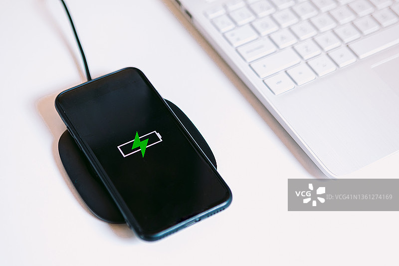 黑色的智能手机，屏幕上有一个绿色闪电的电池标志，在白色的工作台上，在笔记本电脑旁边的充电器底座上充电，没有数据线图片素材