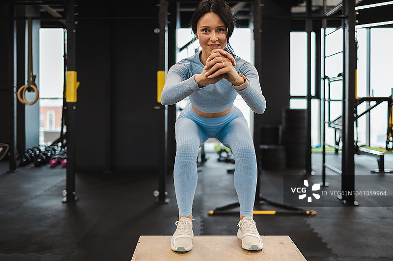 前视图的运动妇女在蓝色运动服做蹲，而站在木制混合健身箱在健身房图片素材