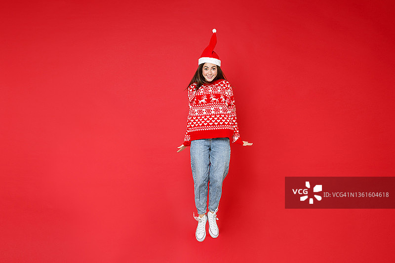 满脸微笑的年轻黑发的圣诞妇女20多岁穿毛衣，圣诞帽跳有乐趣孤立在红色的背景，工作室肖像。祝新年庆佳节快乐理念。图片素材