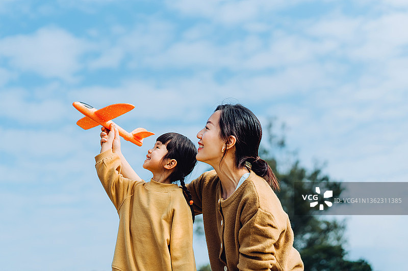 年轻的亚洲母亲和可爱的小女儿一起在户外玩耍，在一个阳光明媚的日子里，在美丽的蓝天下，在公园里开心地笑着，玩着飞机玩具图片素材