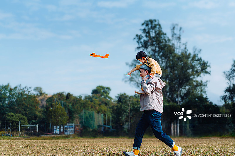快乐的年轻亚洲父亲扛着他的小女儿，花时间在户外，在一个可爱的阳光明媚的日子里，在美丽的蓝天下玩飞机玩具图片素材
