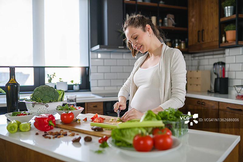 年轻的孕妇在家里用大量的蔬菜准备健康的食物，并使用智能手机寻找一些食谱。健康的饮食习惯图片素材