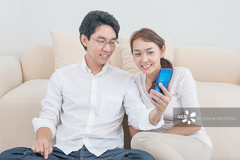 年轻夫妇坐在家里客厅的沙发上用智能手机看在线内容。夫妇在室内看智能手机屏幕。图片素材