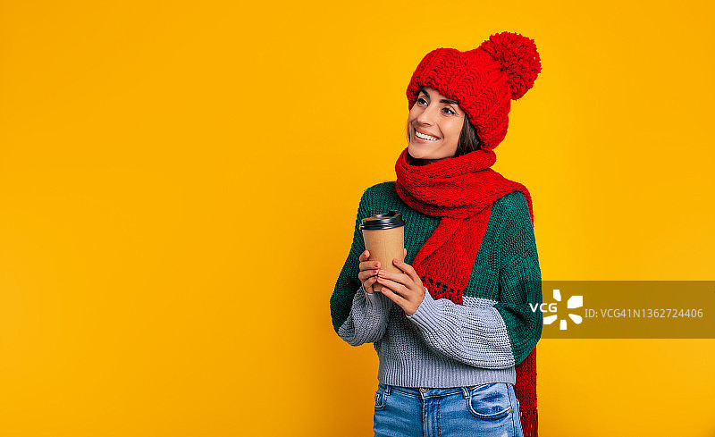 近景肖像快乐的现代黑发女人在红色的帽子和围巾，而她摆姿势与一些饮料在黄色的墙上在工作室图片素材